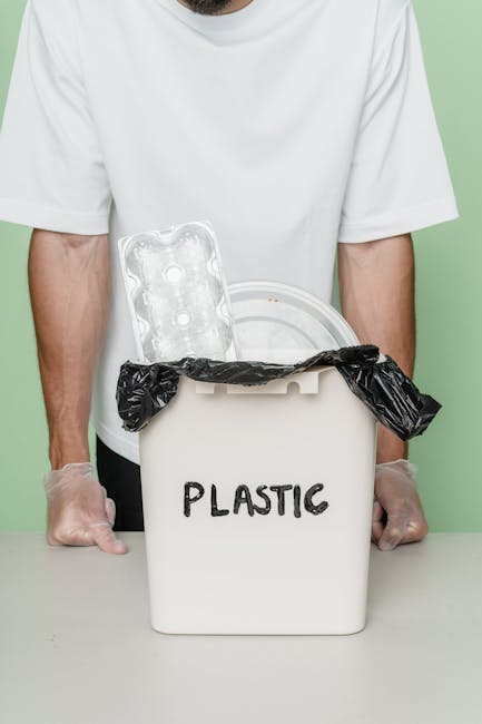 A person wearing a white t-shirt and transparent gloves is holding a white plastic rubbish bin with the word 'PLASTIC' written on it. The bin's lid is open, revealing a black plastic bag lining the interior. On top of the bin, there is a clear plastic egg carton. The background is a plain light green wall, and the scene is set indoors on a grey surface, emphasizing plastic waste disposal and collection practices in environmentally conscious contexts. This visual relates to alternative waste handling options such as separate plastic recycling or disposal that independent rubbish removal services like Rubbish Collection Lewisham may facilitate, especially in areas like Brockley estate where local options are considered.