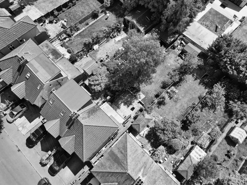 Aerial black-and-white photograph of a residential area showing multiple houses with pitched roofs, some with tile finishes, aligned closely along a street with parked cars in driveway spaces. Behind the row of houses, there is a large, open backyard with a variety of trees casting shadows on the grass, which appears well-maintained. The backyard features a small garden shed, garden beds, and fences dividing different sections. The environment looks tidy, with no visible rubbish or debris, reflecting a scene that could be associated with private waste handling or on-site clearance by a waste management service like Rubbish Collection Lewisham, which specializes in rubbish removal for such properties. The lighting suggests daylight conditions, and the overall scene emphasizes organized, residential waste management and property tidiness.