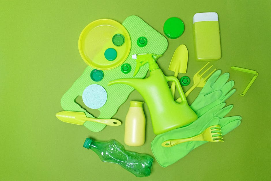A collection of various cleaning and gardening tools and supplies, all predominantly in bright green and yellow tones, arranged on a vibrant green background. The objects include a yellow plastic watering can with a curved handle and long spout, a yellow gardening trowel positioned with its handle angled to the left, and a pair of green rubber gloves placed side by side, fingers pointing to the right. Nearby is a small, yellow plastic bottle, likely for liquid solutions. Also visible are two circular scrub pads, one larger with a textured surface and a smaller solid-colored one, along with a yellow rectangular sponge with a white scrubbing side on the top edge. A green plastic bottle with a screw-on cap lies horizontally at the bottom of the arrangement. Additionally, a small green-handled garden fork with four prongs, and a yellow garden tool with a thin, angled head are present. A green spray bottle with a white nozzle is positioned towards the top right. The scene captures a neatly organized display of horticultural and cleaning tools, suitable for private garden or outdoor work, reflecting a focus on alternative waste handling or rubbish removal services in Lewisham offered by Rubbish Collection Lewisham.