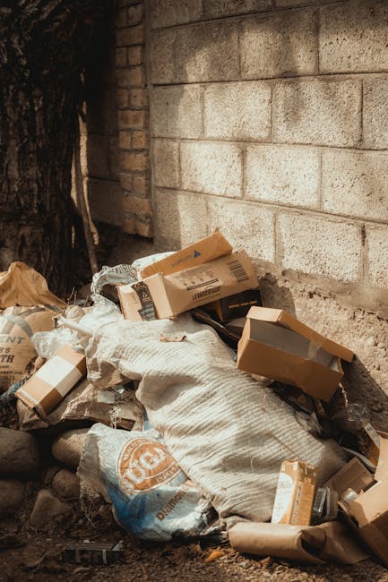 The image shows a red metal skip with a weathered and slightly rusty surface placed between two grey concrete walls. The skip is situated on a dark grey, textured paved surface, with the left wall being smooth and the right wall having a rougher, slightly lighter finish. Next to the skip, there are several transparent plastic bags filled with mixed household waste, including plastic bottles and packaging, some of which are slightly crushed or wrinkled. The scene appears to be outdoors, possibly in an alleyway or a narrow side passage, where space is limited for rubbish disposal. The lighting is neutral, indicating a cloudy day or shaded area, with no direct sunlight or shadows observable. This setting exemplifies an alternative waste handling situation, where individual or private collection services, such as those provided by Rubbish Collection Lewisham, are used to address access issues or tight space constraints for rubbish removal.