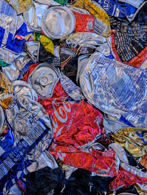 A close-up view of a pile of crushed and flattened aluminium cans, predominantly silver with some featuring red, blue, yellow, and black printed branding. The cans are tightly compacted together, exhibiting wrinkled, crinkled textures typical of aluminum after compression. Around the cans, there are irregular folds and creases, with some cans partially overlapping others, creating a layered appearance. The background shows a mixture of other crushed beverage containers, possibly made of plastic or metal, contributing to a cluttered, waste-filled scene. The overall feel of the image suggests collection or disposal of recyclable waste, which could be part of an alternative waste handling process operated by companies such as Rubbish Collection Lewisham, reflecting the context of independent rubbish removal and recycling services. The lighting is neutral and illuminates the metallic surfaces, highlighting their reflective finishes. This detailed depiction emphasizes the importance of proper waste separation and disposal within the broader scope of rubbish removal services.