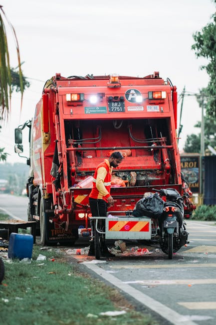 A worker wearing a red and yellow high-visibility vest is operating a large red rubbish collection truck parked on the side of a street, with the rear hatch open as they load waste into the vehicle. The truck, branded with various labels and warning decals, is positioned with its rear close to the pavement, allowing for the efficient collection of rubbish. In front of the truck, a black motorcycle is parked on the sidewalk, with a black plastic garbage bag placed on the motorcycle's seat or rear section. The surrounds include a grassy verge with some scattered litter and a blue container lying on the ground nearby. In the background, a busy roadside scene is visible, with trees and utility poles lining the street, and a small shop or café, along with other vehicles passing by. The lighting is natural, suggesting daytime, with an overcast sky providing diffuse illumination. This scene exemplifies alternative waste handling methods, such as private rubbish collection services, involving on-site loading and disposal outside standard municipal systems, highlighting the importance of efficient rubbish removal for urban and residential areas like Lewisham.