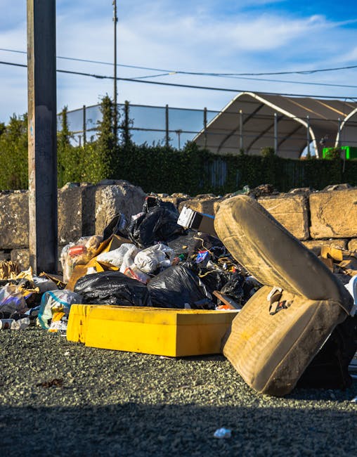 A collection of discarded rubbish piling up on a gravel surface beside a stone wall, primarily consisting of black plastic bin bags filled with waste, with some bags torn open revealing miscellaneous rubble, paper, and packaging materials. In the foreground, an old, stained, beige car seat with fabric upholstery is partially overturned, leaning against the pile of rubbish. Behind the debris, a tall metal pole with an attached sign is visible on the left side of the image, and a chain-link fence topped with dense green foliage extends across the background. To the right, a large, curved, metallic structure — possibly a storage shed or temporary shelter — is partially visible, with the sky above showing a bright, partly cloudy atmosphere typical of an outdoor, urban environment. The scene suggests a context of unattended waste accumulation, highlighting the need for efficient rubbish collection services such as those offered by Rubbish Collection Lewisham, especially for private or alternative waste disposal arrangements.