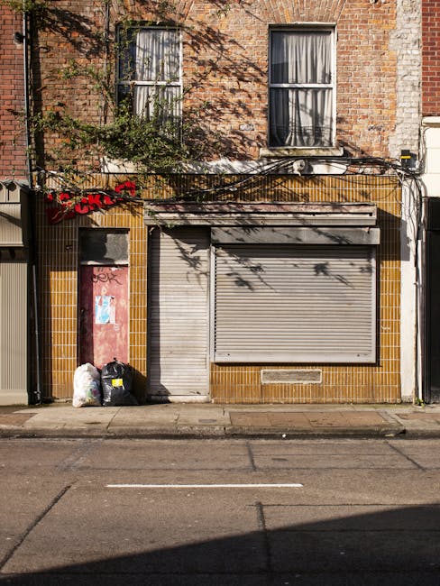 The image depicts the front of a small commercial or retail unit on a city street, with a closed metal roller shutter painted in a light grey tone covering the storefront, which appears weathered and slightly dirty. To the left of the shutter, there is a narrow vertical door painted dusty pink, also showing signs of weathering and graffiti marks. Adjacent to the door, there are two black garbage bags and a white plastic bag placed directly on the pavement, indicating a small rubbish disposal area. The building facade is finished with rectangular yellowish-brown tiles framing the storefront, contrasting with the red brick upper section of the building that features two windows with black frames and partially drawn curtains. Overhead, a network of black cables runs horizontally, casting shadows on the brick wall, with some greenery from a nearby plant or tree partially in view at the top left corner. The pavement in front is made of asphalt with a single white line marking a parking space. This scene, typical of urban environment with cluttered rubbish, subtly relates to independent waste collection solutions offered by companies such as Rubbish Collection Lewisham, providing alternative options for traders and shop owners seeking private disposal or on-site clearance services.