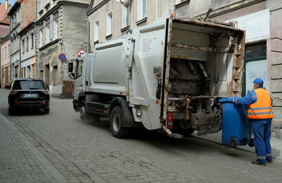 A waste collection truck parked on a narrow cobblestone street in an urban residential area with older, multi-story buildings lining the roadside. The truck is white with visible rust and dirt on its metal rear section, which is open to reveal mechanical components used for compacting waste. A worker dressed in a blue uniform and an orange high-visibility vest is standing beside the truck, holding a blue wheeled trash bin and preparing to load it into the vehicle. To the left, there is a black car parked parallel to the curb, and various road signs indicating parking restrictions are mounted on building facades. The scene is lit with soft natural daylight, and the environment suggests an alternative waste collection or on-site rubbish removal service, such as that offered by Rubbish Collection Lewisham, operating within an inner-city setting.