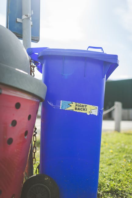 A tall, bright blue rubbish bin with a hinged lid, positioned on a grassy outdoor area adjacent to a pink waste container with black circular ventilation holes and a gray lid, both linked by a chain. The blue bin has a yellow and blue sticker with the words 'Fight Back!' partially torn, and is situated near a pole with a sign in the background. The scene is illuminated by natural daylight, with a partly cloudy sky and blurred background elements including a road barrier, suggesting an outdoor setting for private waste management or alternative rubbish removal services, as represented by Rubbish Collection Lewisham.