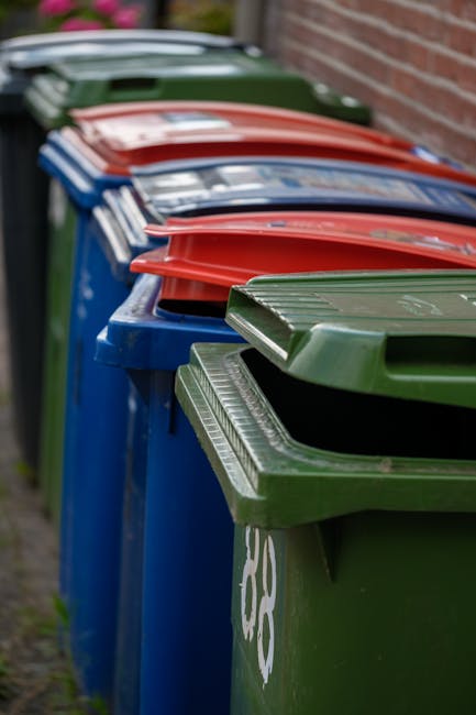 A tall, bright blue rubbish bin with a hinged lid, positioned on a grassy outdoor area adjacent to a pink waste container with black circular ventilation holes and a gray lid, both linked by a chain. The blue bin has a yellow and blue sticker with the words 'Fight Back!' partially torn, and is situated near a pole with a sign in the background. The scene is illuminated by natural daylight, with a partly cloudy sky and blurred background elements including a road barrier, suggesting an outdoor setting for private waste management or alternative rubbish removal services, as represented by Rubbish Collection Lewisham.
