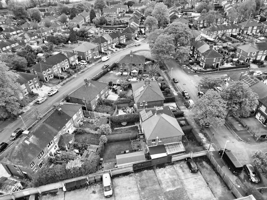 An aerial view of a residential neighbourhood showing multiple detached and semi-detached houses with pitched roofs, garden areas, and driveways. The foreground features a row of houses with backyards separated by fences and hedges, some with sheds, garden furniture, and paving slabs. A paved street runs through the area, with several parked cars and a few moving vehicles visible. Mature trees with full canopies are scattered throughout the area, providing shade and greenery. The background displays additional rows of houses and streets extending into the distance, with rooftops varying in material and color. The overall scene is well-lit with natural daylight, emphasizing the suburban density and the arrangement of individual properties within the neighbourhood. This detailed description supports visual understanding of typical residential waste management and the importance of proper rubbish collection in such environments, aligning with services like those offered by Rubbish Collection Lewisham for private waste handling and on-site clearance.