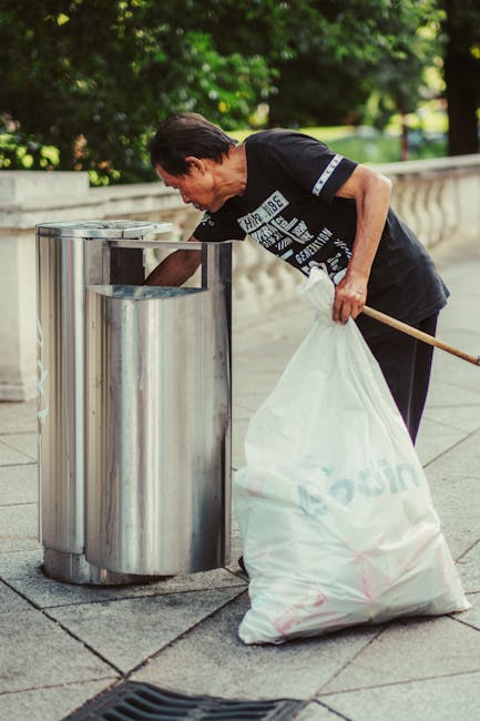 A middle-aged woman with short dark hair, wearing a black t-shirt and dark pants, is engaged in rubbish collection on a paved sidewalk. She is holding a large white plastic trash bag in her left hand, which appears to be partially filled with waste, and is leaning forward slightly to deposit or retrieve rubbish from a cylindrical stainless steel trash bin with a shiny, reflective surface. The trash bin is positioned against a low stone wall, with lush green foliage and trees in the background, suggesting an outdoor urban environment. The scene is well-lit with natural daylight, emphasizing the materials and textures of the trash bag, the metallic surface of the bin, and the paving stones beneath her feet. The activity illustrates a focus on private or informal waste disposal, aligning with the services offered by Rubbish Collection Lewisham for rubbish removal and free-standing on-site clearance.