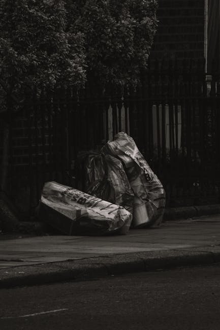 Three large black plastic garbage bags, partially crumpled and tied at the top, are placed on a concrete sidewalk in front of a black metal fence. The bags appear to contain waste and are situated near the kerb, with a dark, shadowed background that includes some foliage from trees or bushes behind the fence. The scene likely depicts an area awaiting rubbish collection, reflecting typical private waste handling or alternative rubbish removal options. The environment is urban, with the bags positioned on a paved walkway beside a road, under dim lighting conditions, possibly dusk or overcast weather. The image emphasizes the importance of proper waste disposal, relating to services such as those offered by Rubbish Collection Lewisham, for effective rubbish removal and on-site clearance.