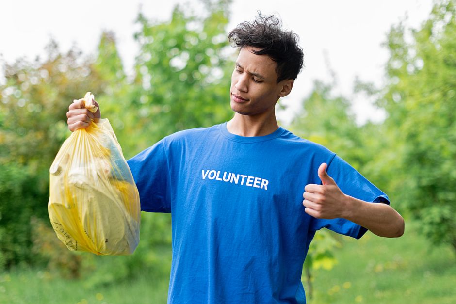 A young male volunteer with curly dark hair is standing outdoors in a green, leafy environment during daytime. He is wearing a bright blue T-shirt with the word 'VOLUNTEER' printed in white across the chest. He holds a yellow plastic bag filled with collected waste in his right hand, gripping the top of the bag firmly. His facial expression suggests focus or mild concern as he looks down at the bag. The background features blurred green foliage, with natural light illuminating the scene evenly. The moment captures the volunteer engaged in community clean-up or rubbish collection activity, emphasizing environmentally friendly efforts and the importance of waste management, which aligns with the theme of rubbish removal services. The scene conveys a sense of civic responsibility and environmental consciousness associated with private waste disposal and rubbish clearance actions.