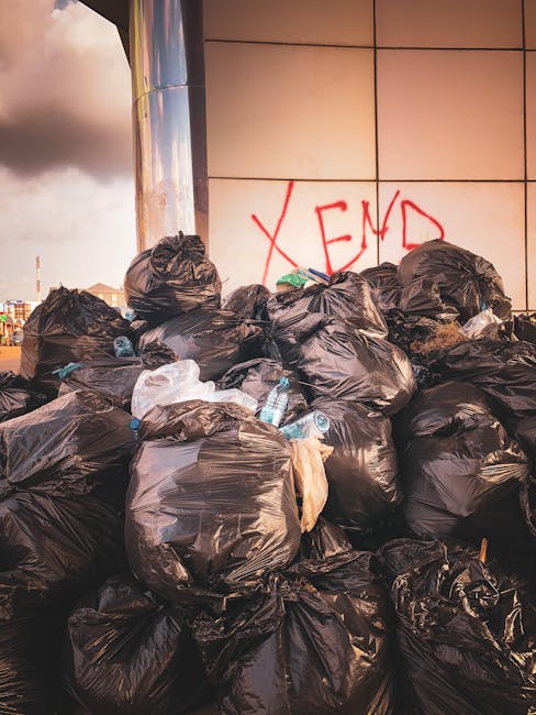 A large pile of black plastic rubbish bags is stacked against a modern building with a tiled exterior wall. The bags are tightly packed, some with visible crumples and creases, and are of varying sizes, indicating different types of waste. Among the bags, clear plastic water bottles and other small items are partially visible, protruding from some of the bags. The area appears to be outdoors, possibly on a paved surface or driveway, with ambient daylight casting soft shadows on the bags. In the background, graffiti with the letters 'XEND' is spray-painted in red on the building's wall. The scene suggests an instance of roadside or site waste accumulation that might be subject to private collection or alternative rubbish removal services, such as those provided by Rubbish Collection Lewisham, rather than official municipal waste disposal. This visual representation underscores the importance of professional rubbish removal to manage and clear waste efficiently in urban environments.